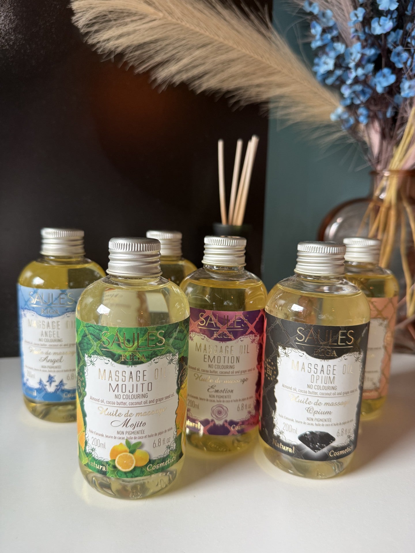 assortiment van luxe Saules Fabrika massageoliën in 200ml doseerflessen, waaronder Mojito, Emotion en Opium, gepresenteerd op een witte tafel met een sfeervolle achtergrond van droogbloemen en geurstokjes - Glow Mystic in Style.