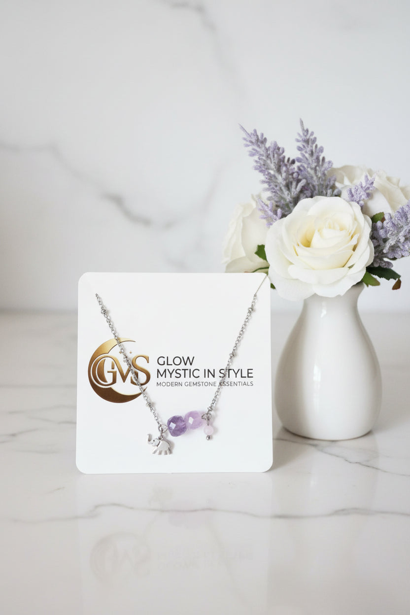 Elegant zilveren armbandje met facetgeslepen paarse amethist kralen en een geluksolifantje op een wit displaykaartje van Glow Mystic in Style, gefotografeerd naast een vaasje met een witte roos.