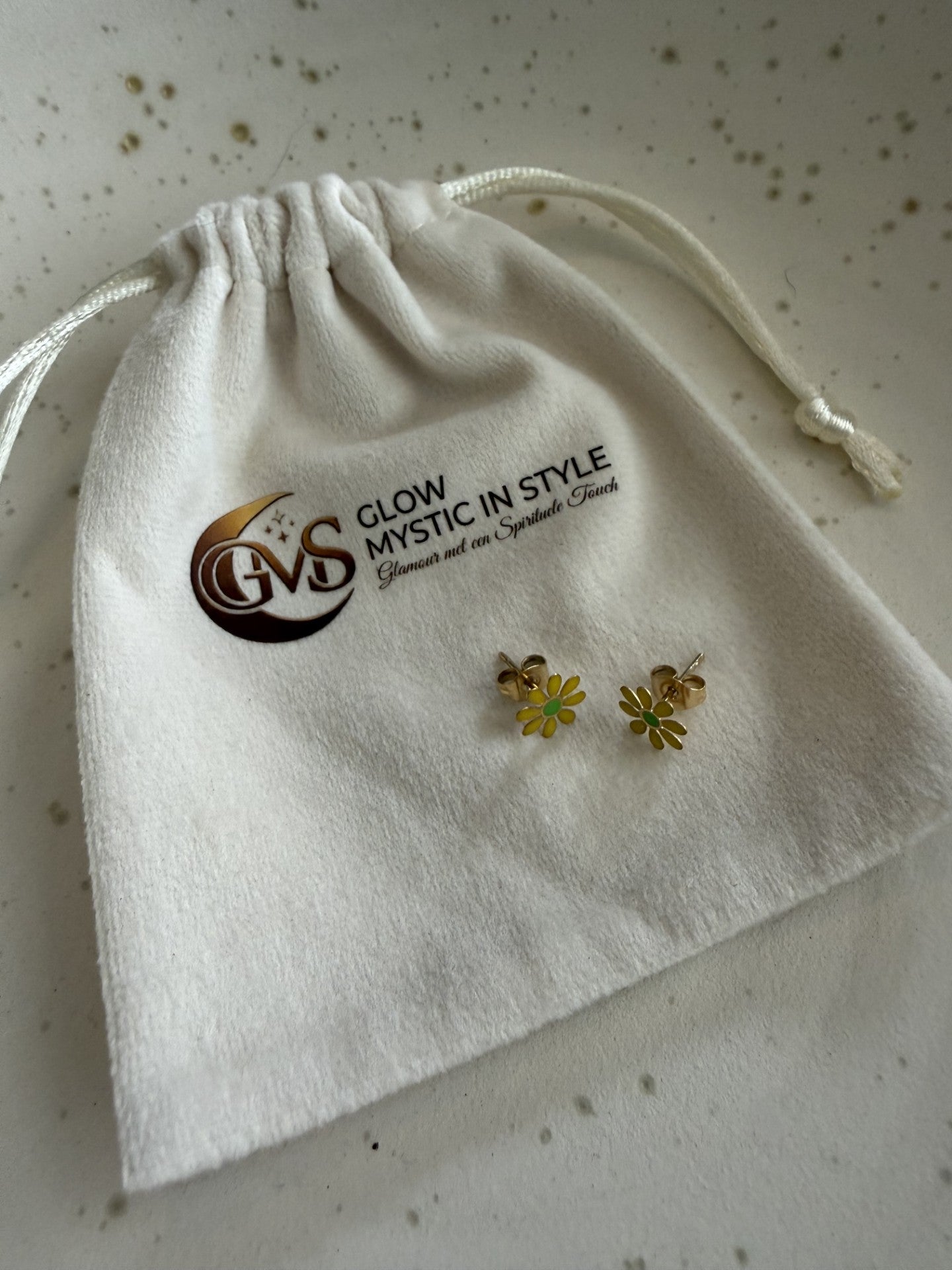 "Productfoto van Glow Mystic in Style gele bloem-oorknopjes. De oorbellen rusten tegen een luxe, crème-wit fluwelen zakje waarop het goudkleurige logo van Glow Mystic in Style prijkt. Het geheel ligt op een lichte marmeren ondergrond met zachte grijze aders, wat een elegante en professionele uitstraling geeft aan de verpakking en het sieraad."