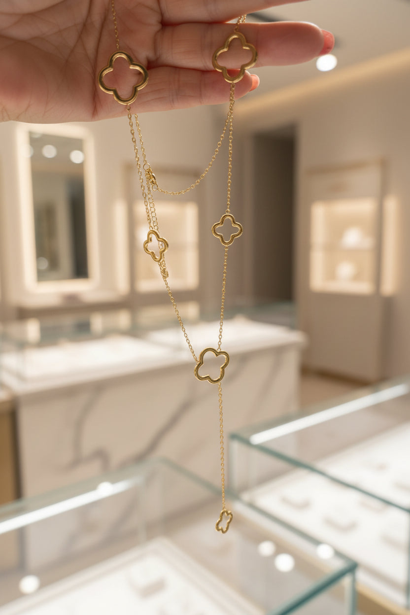 Een goudkleurige , gelaagde ketting met meerdere open klavertjevier elementen van verschillende groottes, omhooggehouden in een hand tegen de lichte, wazige achtergrond van een luxe sieradenboetiek met verlichte vitrines.