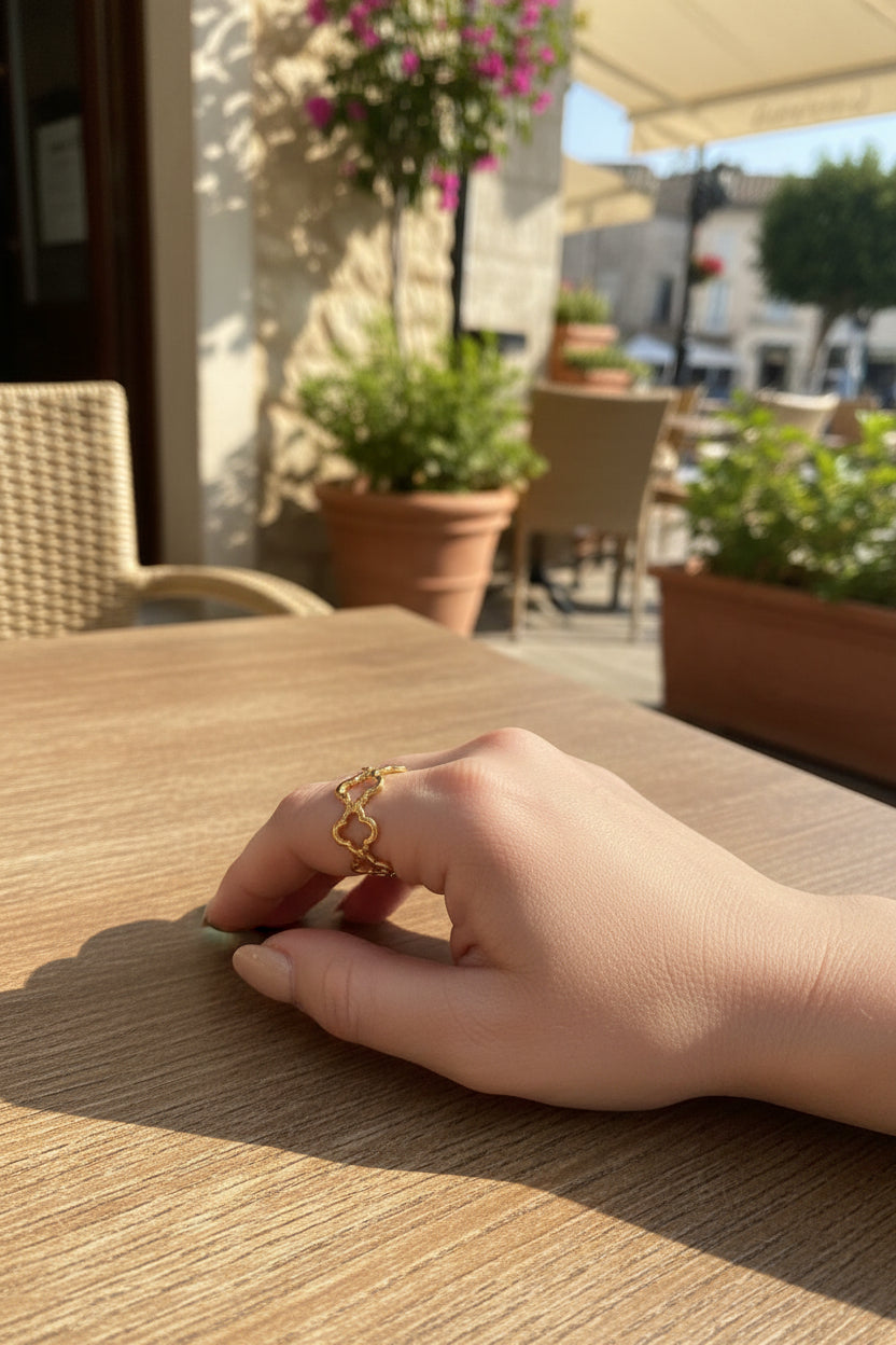 "Goudkleurige RVS klaver ring gedragen aan de hand tegen een achtergrond van een tafel op een terrasje in de zon met potten met planten eromheen.