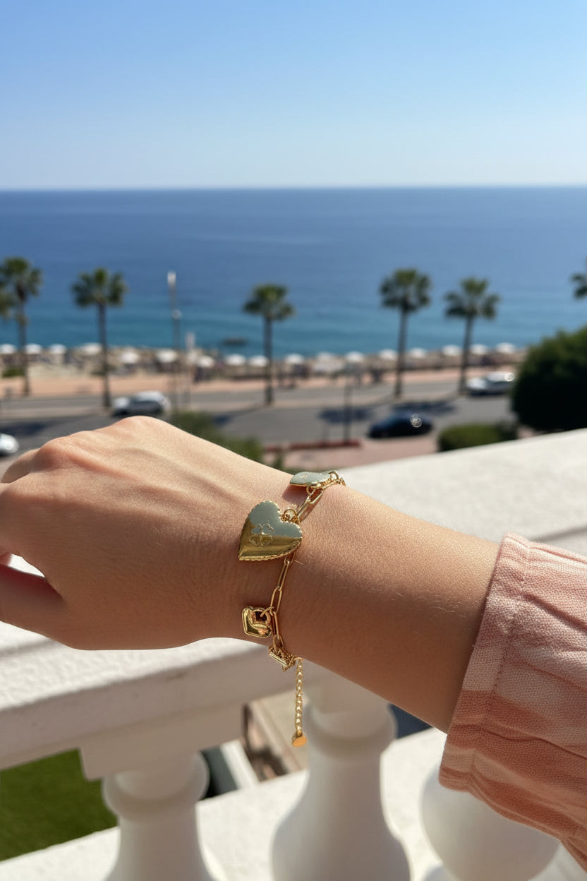 Close-up van een hand die een gouden bedelarmband met hartjes draagt, gefotografeerd vanaf een wit balkon met p de achtergrond een zonnig uitzicht op de blauwe zee en palmbomen.