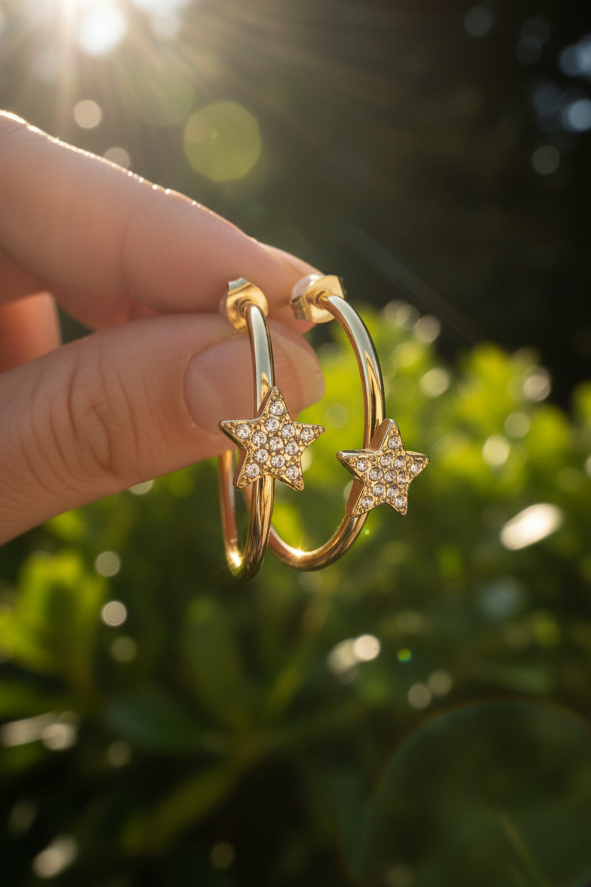 gouden stainless steel creolen met fonkelende strass sterren tussen de vingers in de natuur met zonlicht die op de oorbellen schijnt.