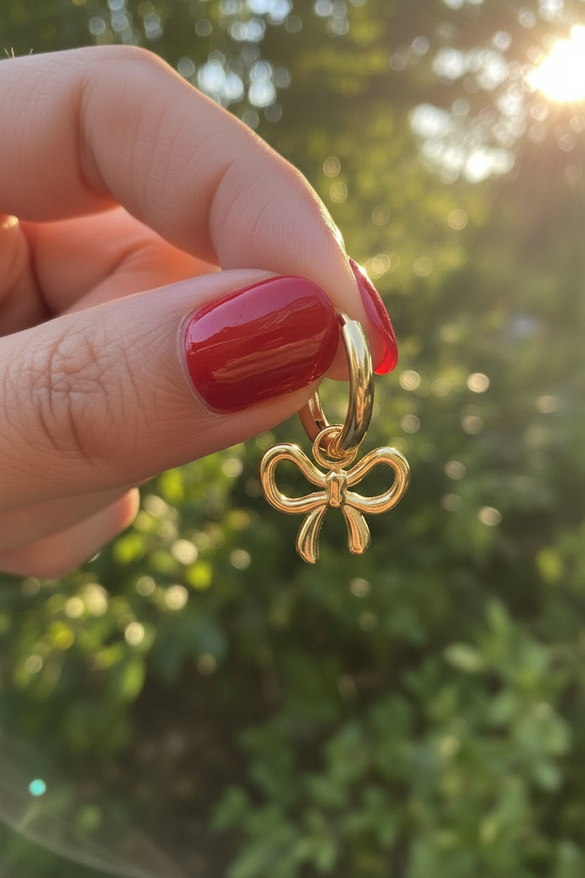 Trendy gouden waterproof RVS bow hoop oorbellen met strikje, vastgehouden in de hand in de natuur met sfeervol, warm zonlicht en een luxe esthetische uitstraling.