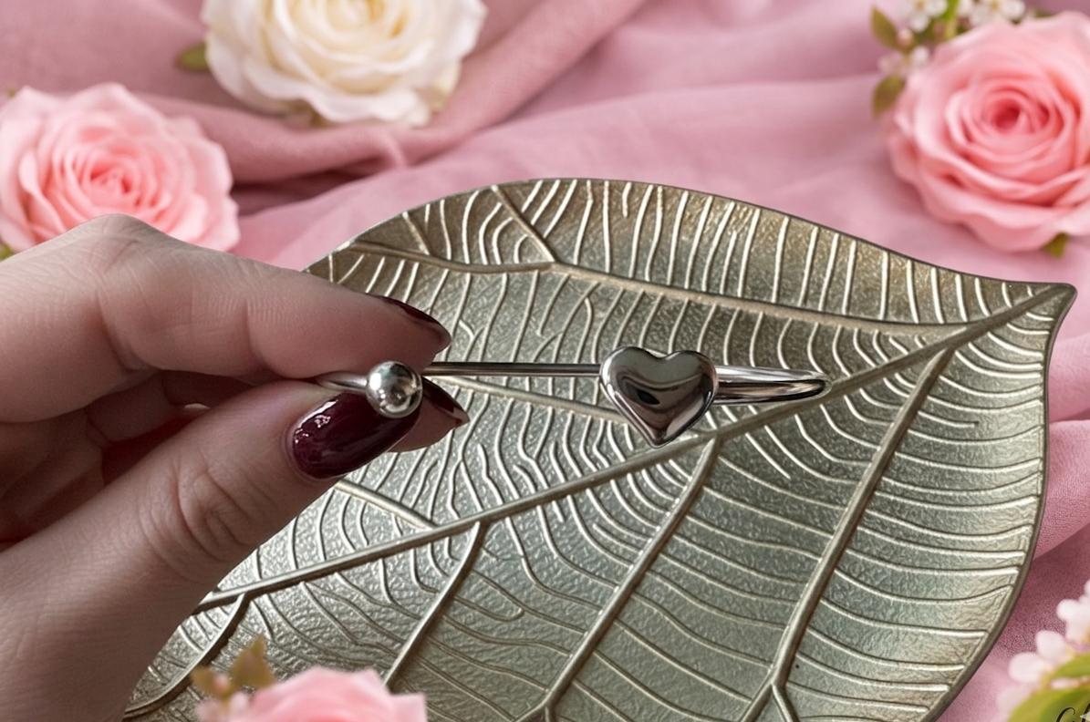 Detailopname van een minimalistische zilveren RVS-bangle met hartje, vastgehouden door een hand met donkerrode, perfect verzorgde nagels. Het sieraad rust op een goudkleurig metalen blaadje tegen een achtergrond van zachtroze stoffen en bloemen. Glow Mystic in Style.