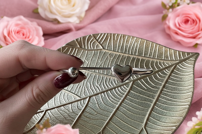 Detailopname van een minimalistische zilveren RVS-bangle met hartje, vastgehouden door een hand met donkerrode, perfect verzorgde nagels. Het sieraad rust op een goudkleurig metalen blaadje tegen een achtergrond van zachtroze stoffen en bloemen. Glow Mystic in Style.