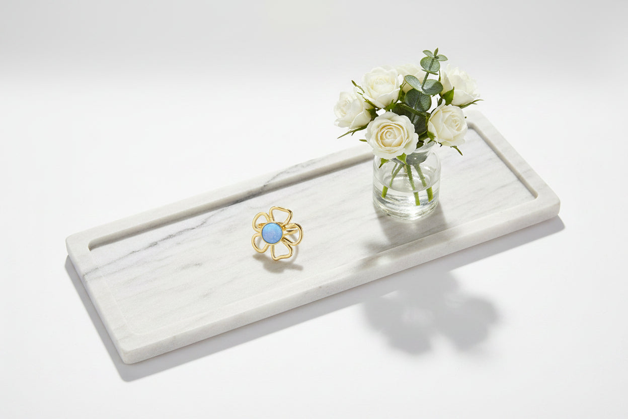 Goudkleurige RVS bloemenring met een ronde, lichtblauwe steen op een wit marmeren blad met witte rozen.