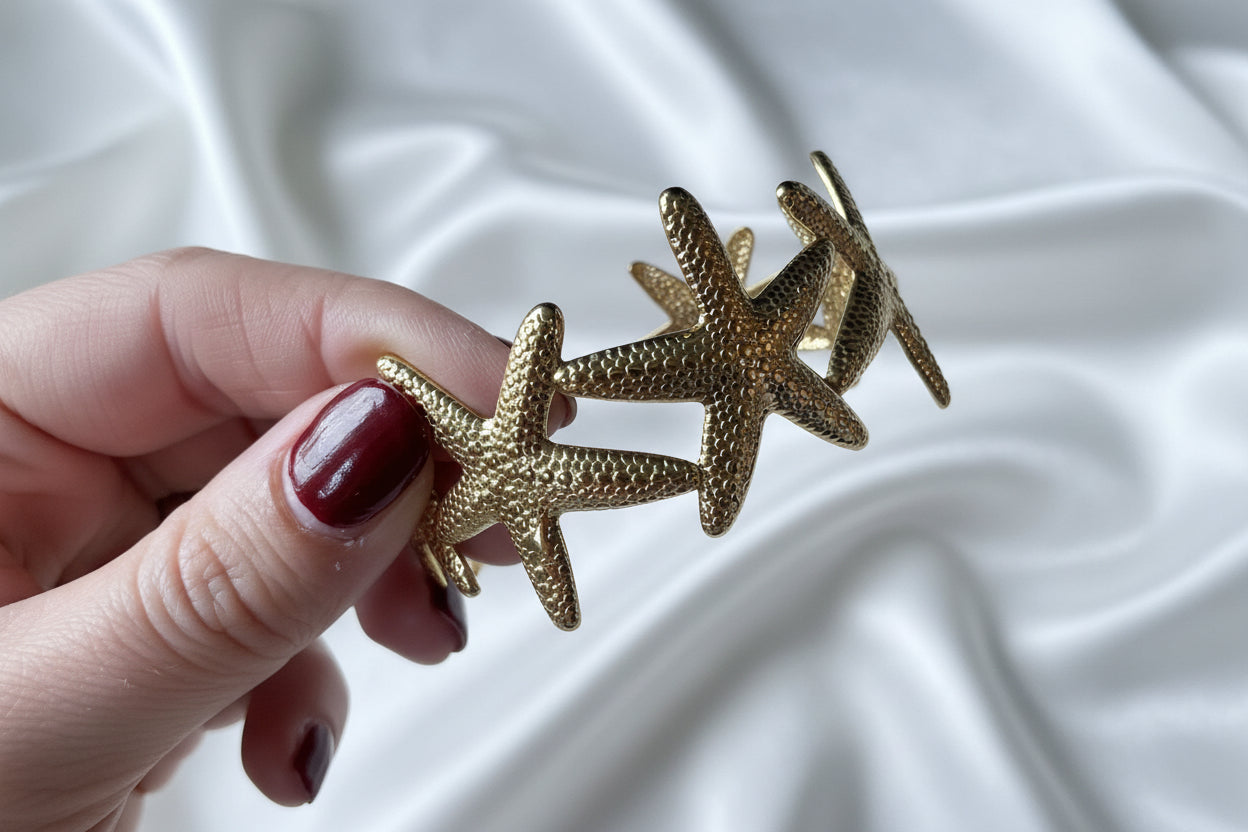 Een hand die de gouden zeester bangle armband vasthoudt boven een wit satijnen achtergrond, wat een duidelijk beeld geeft van de grootte en de luxe afwerking van het sieraad.