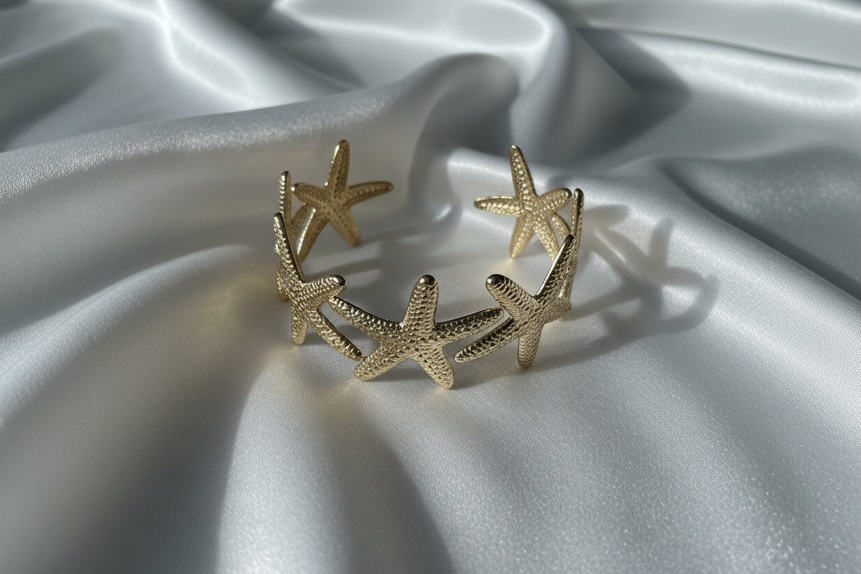 Luxe gouden zeester bangle van stainless steel gepresenteerd op een glanzend wit satijnen kleed, waarbij de details van de zeesterren prachtig uitkomen in het zachte licht.