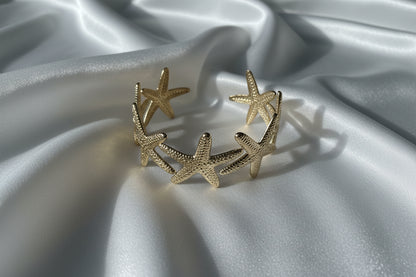 Luxe gouden zeester bangle van stainless steel gepresenteerd op een glanzend wit satijnen kleed, waarbij de details van de zeesterren prachtig uitkomen in het zachte licht.