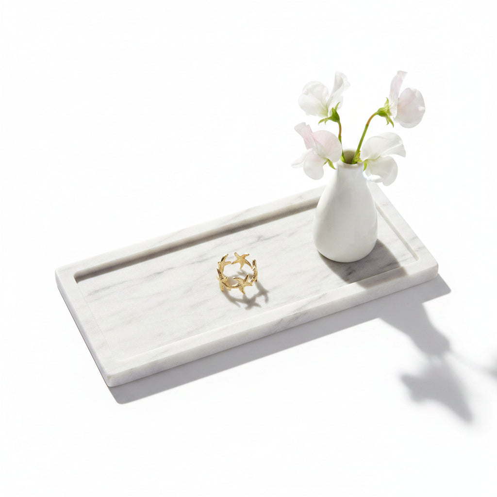 Goudkleurige RVS ring met zeesterren op een rechthoekig marmeren dienblad naast een klein wit vaasje met witte bloemen.