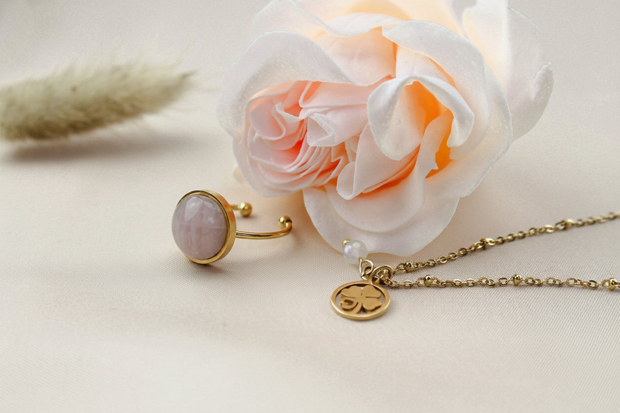 Sfeervolle close-up van een goudkleurige stainless steel ketting met een klavertje vier met hartje bedel en een regenboog maansteen kraal, liggend naast een roze-oranje roos en gedroogd gras op een crèmekleurige satijnen achtergrond. Een bijpassende Maansteen-ring is ook zichtbaar.