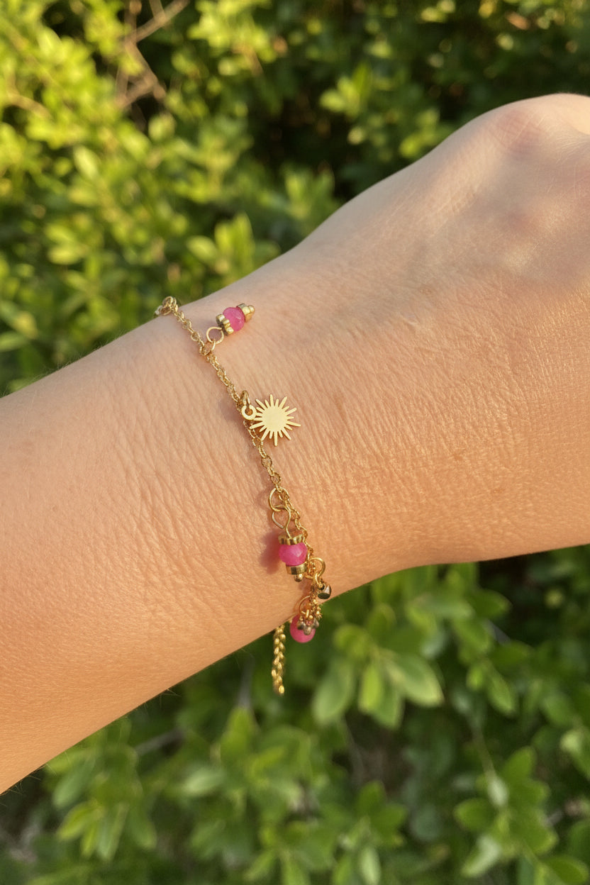 Goudkleurig stainless steel armbandje met een zonnestraal bedel en paars-roze kraaltjes om de arm van een vrouw op een zonnig strand van Glow Mystic in Style.