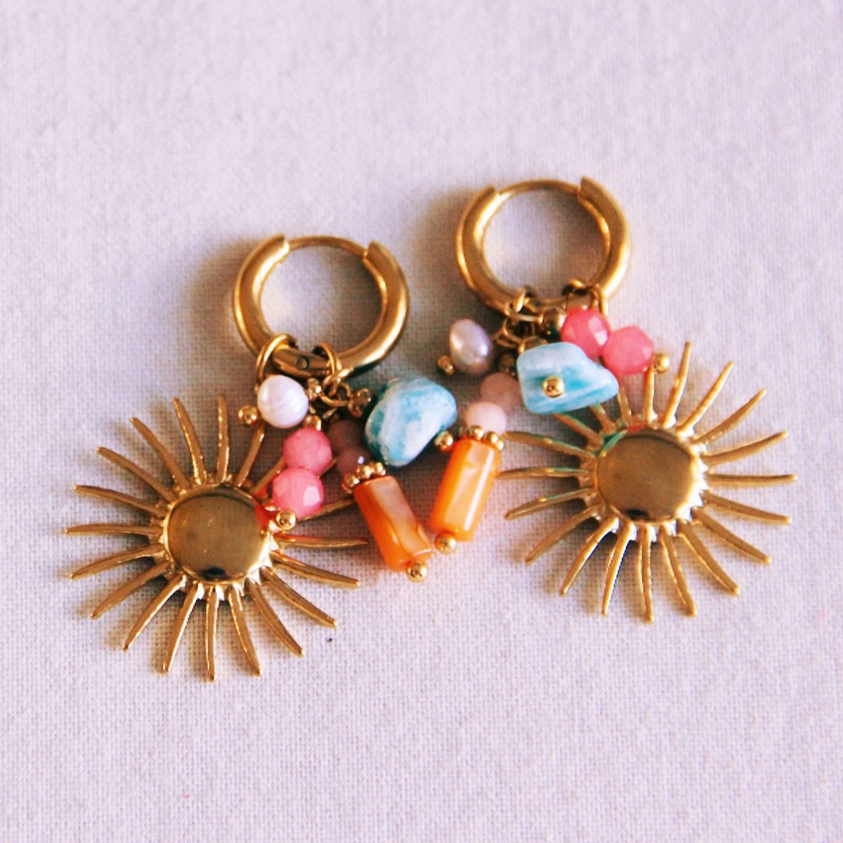 "Close-up van een paar gouden roestvrijstalen hoops met zonhangers, turquoise steentjes, roze facetkralen en oranje bedeltjes."