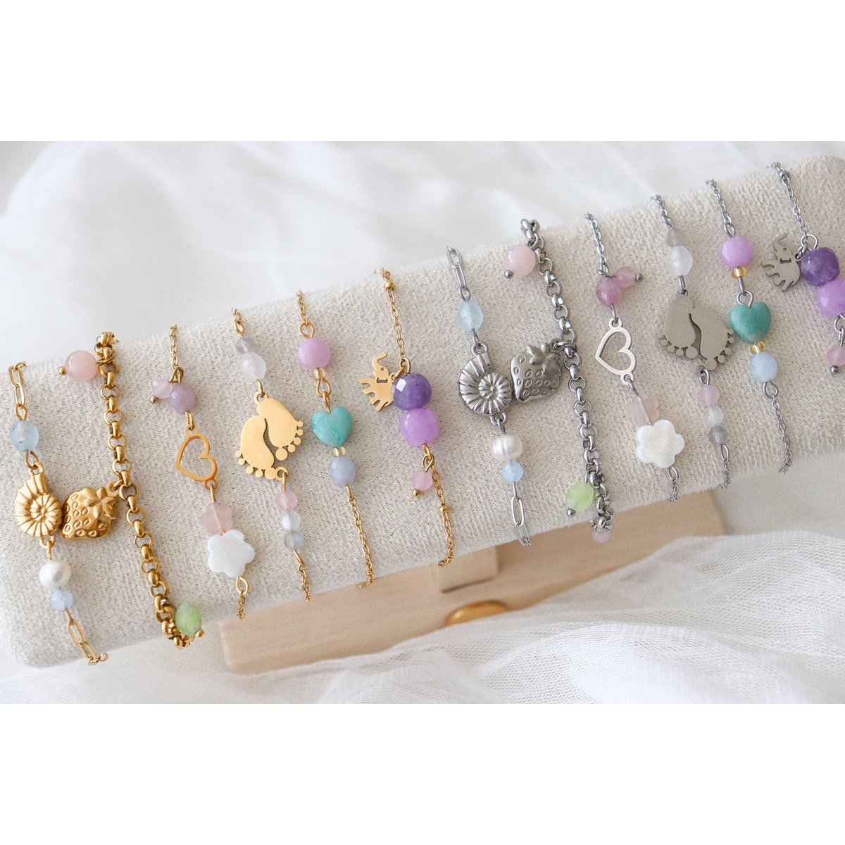 "Bovenaanzicht van het zilverkleurige Saint Clairmont 'Liefdevol Begin' edelsteen armbandje op een stoffen sieradendisplay, met focus op de zilveren babyvoetjes-bedel en de combinatie van Rozenkwarts en Maansteen."