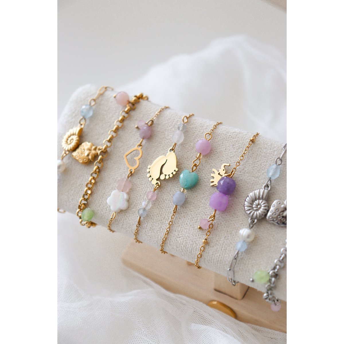 "Bovenaanzicht van het Saint Clairmont 'Liefdevol Begin' edelsteen armbandje op een stoffen sieradendisplay. De focus ligt op de gedetailleerde gouden babyvoetjes-bedel en de delicate combinatie van Rozenkwarts en Maansteen kralen aan de verfijnde schakelketting."
