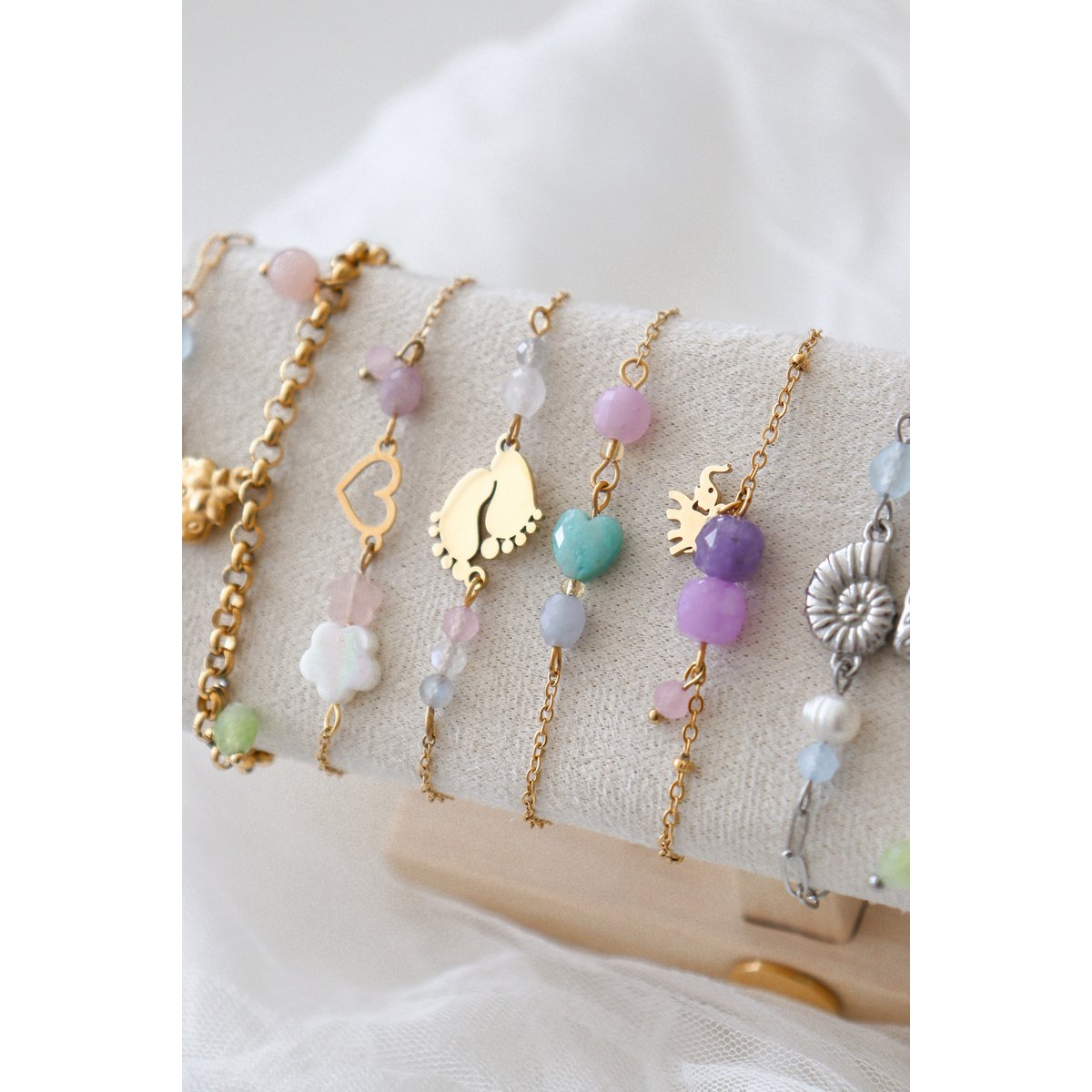 Bovenaanzicht van het Saint Clairmont 'Liefdevol Begin' edelsteen armbandje op een stoffen sieradendisplay. De focus ligt op de gedetailleerde gouden babyvoetjes-bedel en de delicate combinatie van Rozenkwarts en Maansteen kralen aan de verfijnde schakelketting."