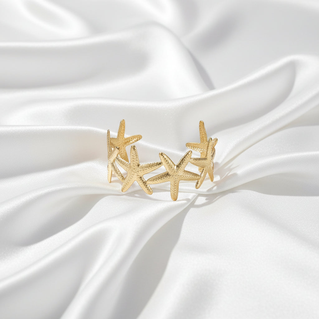 Brede gouden bangle armband van Glow Mystic gemaakt van stainless steel met een gedetailleerd patroon van zeesterren, gefotografeerd op een witte satijnen achtergrond.