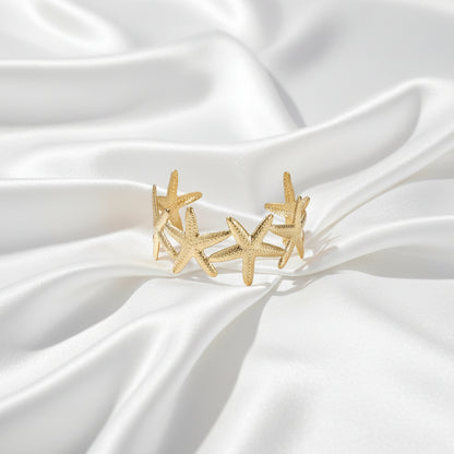 Brede gouden bangle armband van Glow Mystic gemaakt van stainless steel met een gedetailleerd patroon van zeesterren, gefotografeerd op een witte satijnen achtergrond.