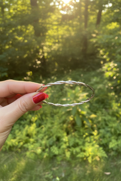Zilveren gedraaide RVS armband subtiel vastgehouden tussen de vingers met nagellak in de natuur met sfeervolle zonnestralen.