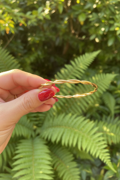 Goudkleurige gedraaide RVS armband subtiel vastgehouden tussen de vingers in de natuur met sfeervol glanzende zonnestralen.