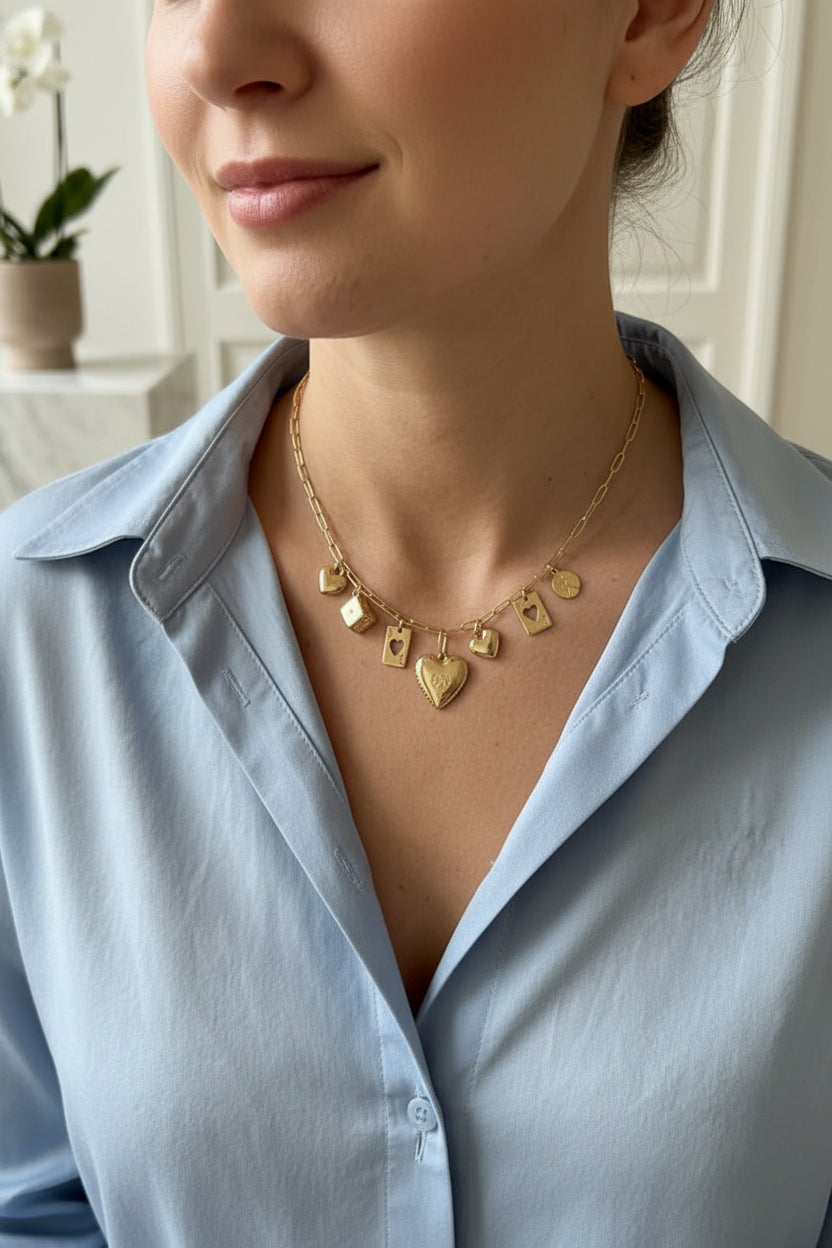 Goudkleurige RVS charmsketting met dobbelsteen-, speelkaarten- en hartbedels gedragen om de hals van een vrouw in een zacht blauwe kleur blouse.