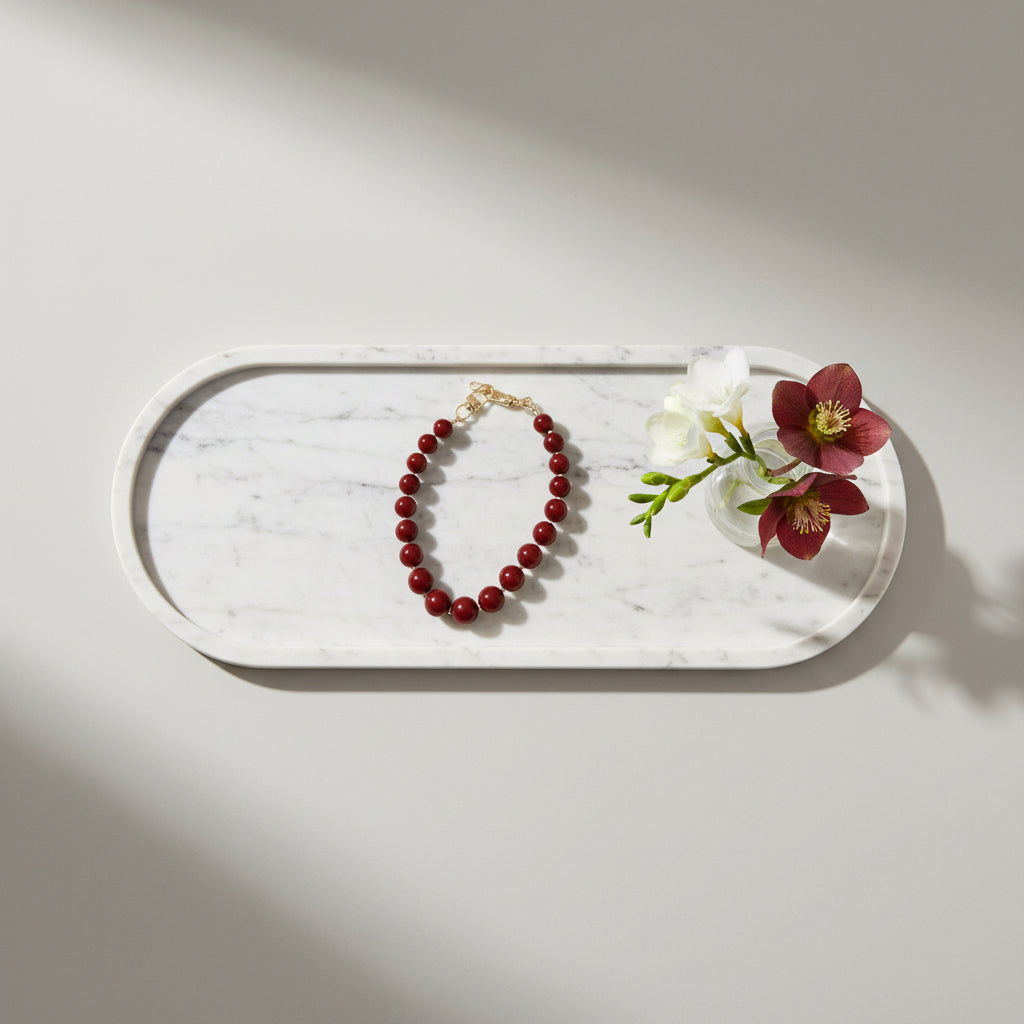 Berry red telefoonkoord met ronde kralen en goudkleurige sluiting, stijlvol gepresenteerd op een wit marmeren dienblad naast decoratieve bloemen.