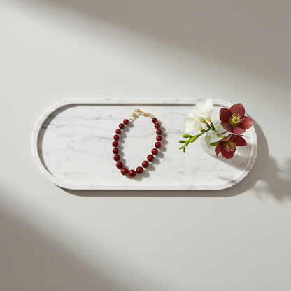 Berry red telefoonkoord met ronde kralen en goudkleurige sluiting, stijlvol gepresenteerd op een wit marmeren dienblad naast decoratieve bloemen.