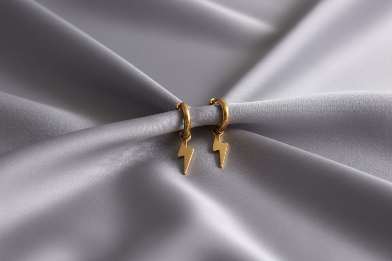 Minimalistische goudkleurige oorbellen met bliksem hanger 12 mm op grijs satijnen kleed