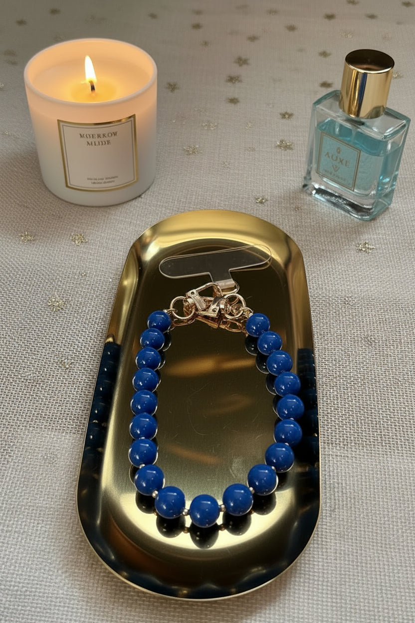 Vooraanzicht van een blauwe kralen telefoonkoord met gouden details op een goudkleurige schaal met lifestyle producten eromheen.