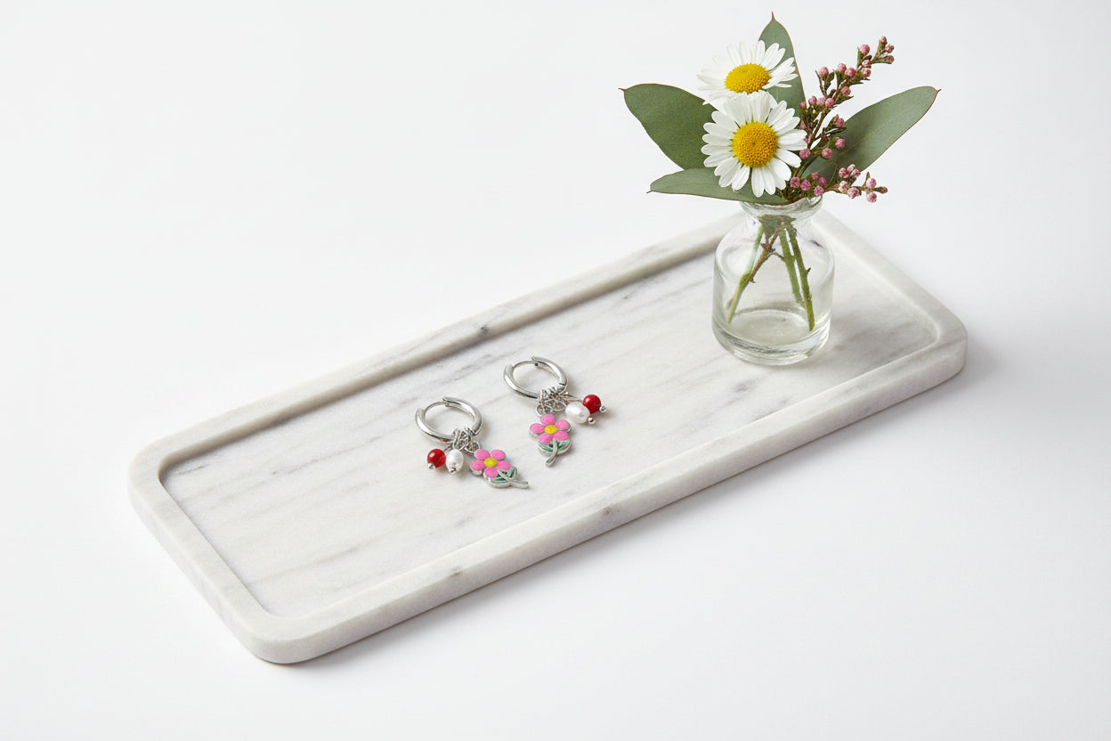 Zilveren hangoorbellen met een roze bloemetje, zoetwaterparel en rood kraaltje, liggend op een langwerpig wit marmeren dienblad naast een glazen vaasje met madeliefjes en eucalyptus.