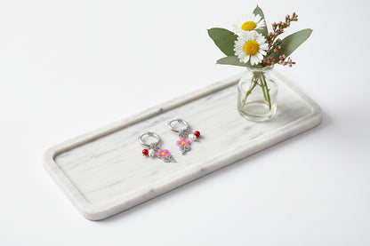 Zilveren hangoorbellen met een roze bloemetje, zoetwaterparel en rood kraaltje, liggend op een langwerpig wit marmeren dienblad naast een glazen vaasje met madeliefjes en eucalyptus.