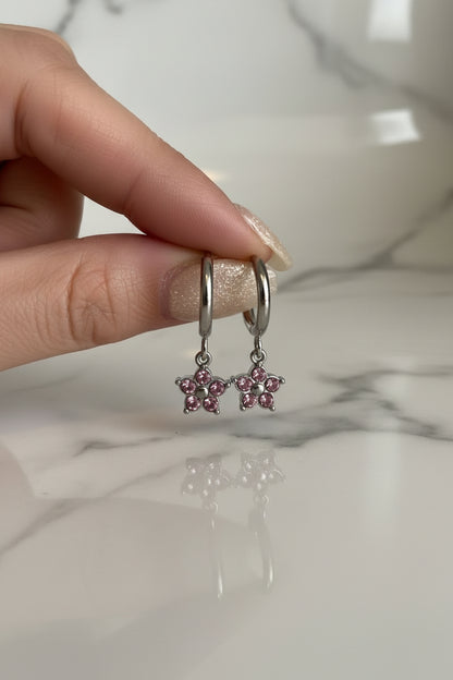 Gedetailleerde close-up van een hand met glitter nagellak die een paar RVS bloem oorbellen met roze kristallen vasthoudt op een marmeren ondergrond.