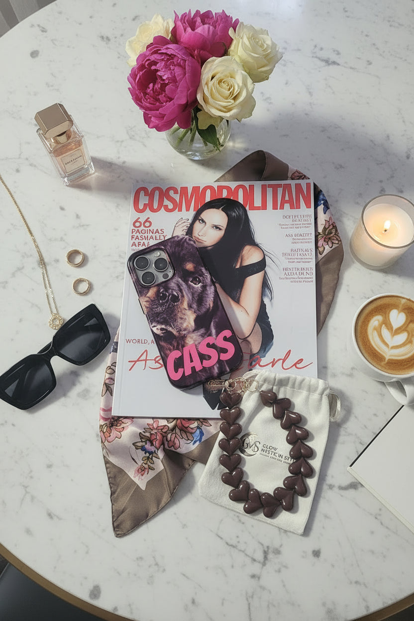 Trendy bruin hartjes telefoonkoord op Cosmopolitan magazine met koffie, bloemen, parfum en smartphone als luxe lifestyle setting.