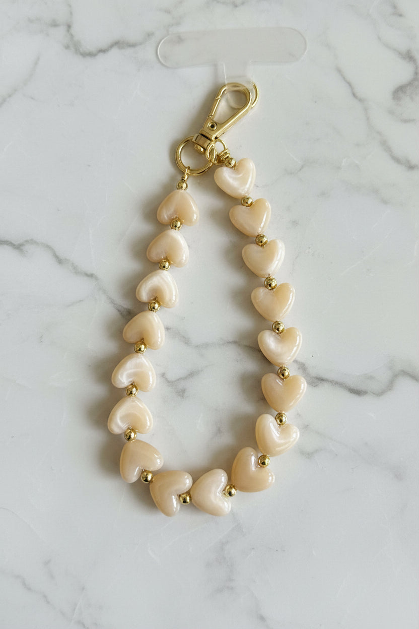 Luxe soft mocha kleur telefoonkoord gemaakt van glanzende parelmoer hartjeskralen en goudkleurige details met een hartjesbedel gepresenteerd op een marmeren achtergrond.