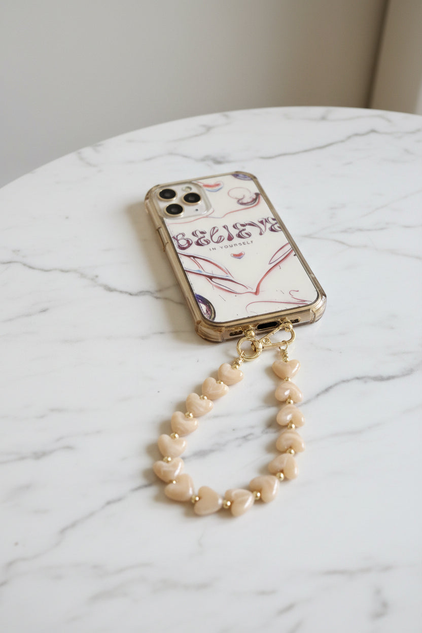 Luxe soft mocha kleur telefoonkoord gemaakt van glanzende parelmoer hartjeskralen en goudkleurige details met een hartjesbedel gepresenteerd op een marmeren achtergrond.
