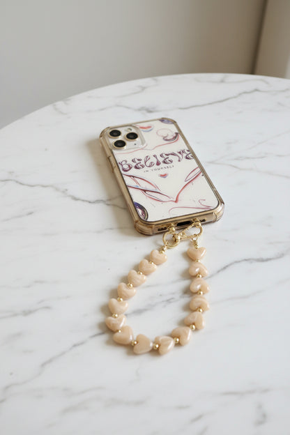Luxe soft mocha kleur telefoonkoord gemaakt van glanzende parelmoer hartjeskralen en goudkleurige details met een hartjesbedel gepresenteerd op een marmeren achtergrond.