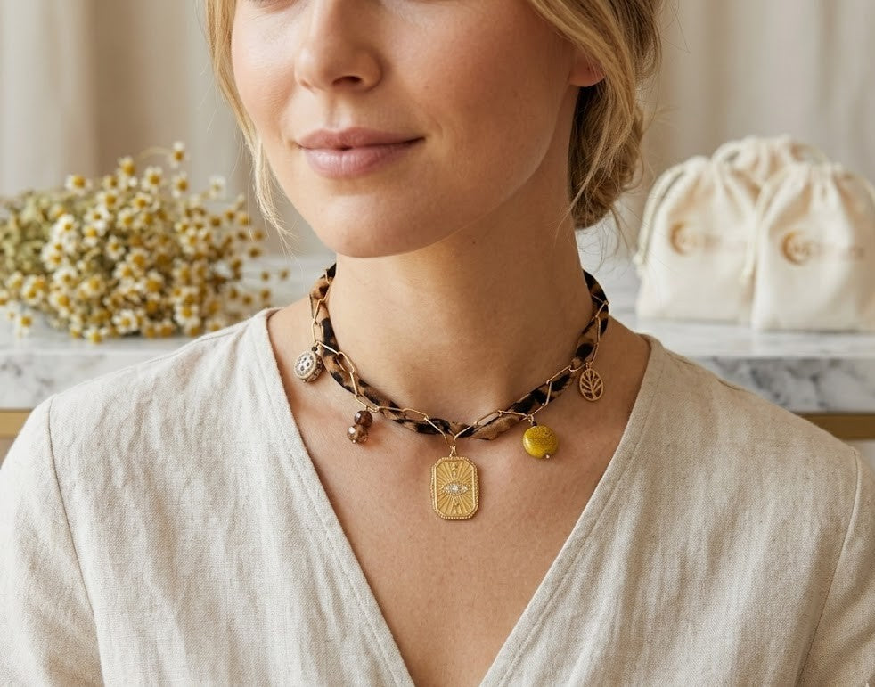 "Close-up van een model dat de handgemaakte panterprint choker van Glow Mystic draagt. De goudkleurige Evil Eye hanger met kristallen vormt het stralende middelpunt. De ketting valt elegant op de hals en wordt getoond tegen een achtergrond van een luxe, zandkleurige blouse in een sfeervolle setting."