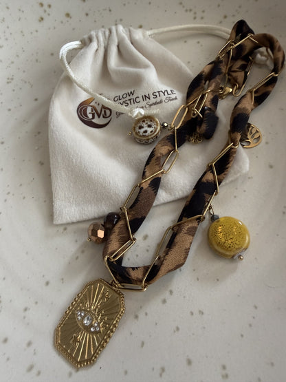 "Bovenaanzicht van de handgemaakte panterprint choker liggend op een witte gespikkelde schaal. Naast de ketting liggen twee luxe crème-witte fluwelen cadeauzakjes met het goudkleurige Glow Mystic in Style logo. De foto toont alle details van de bedels en de RVS schakels."