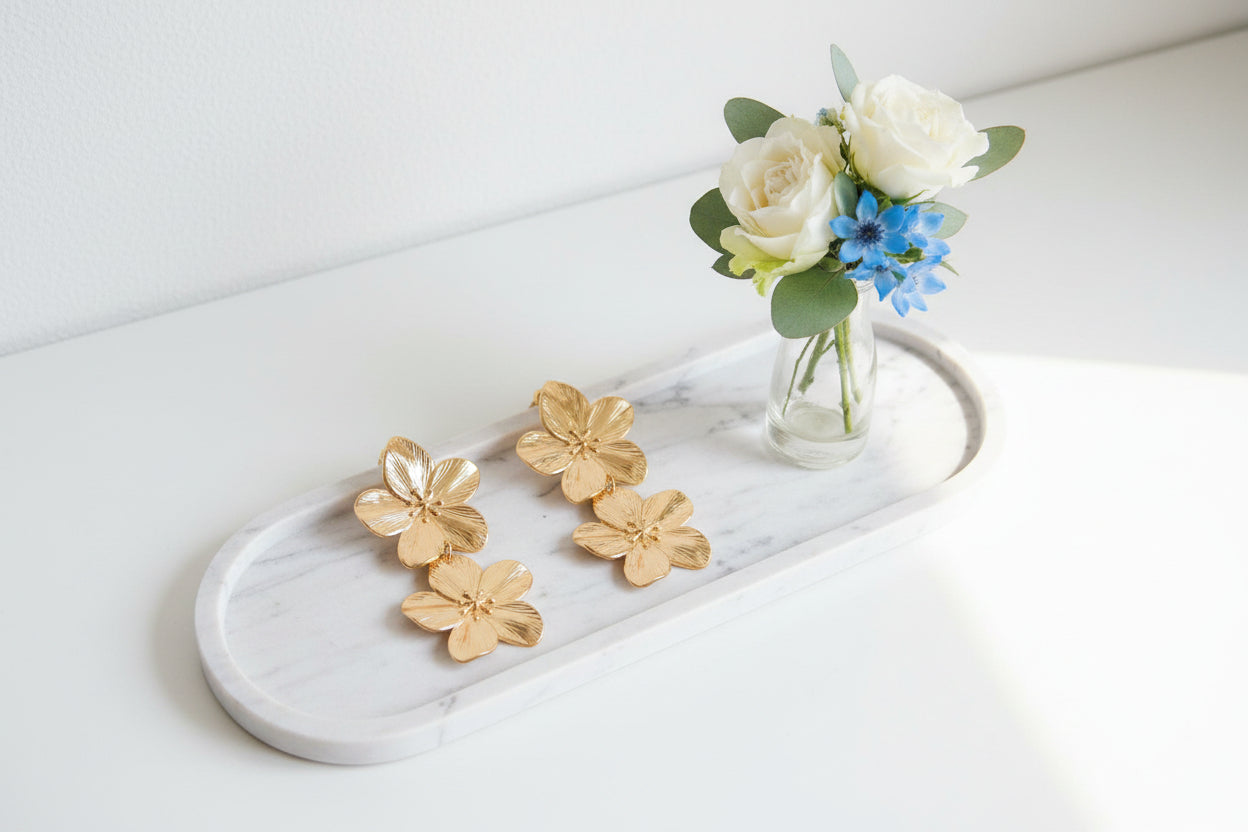Goudkleurige oorbellen dubbele bloem van RVS, lichtgewicht statement damesaccessoire gepresenteerd op een langwerpig marmeren schaal met een vaasje bloemen ernaast.