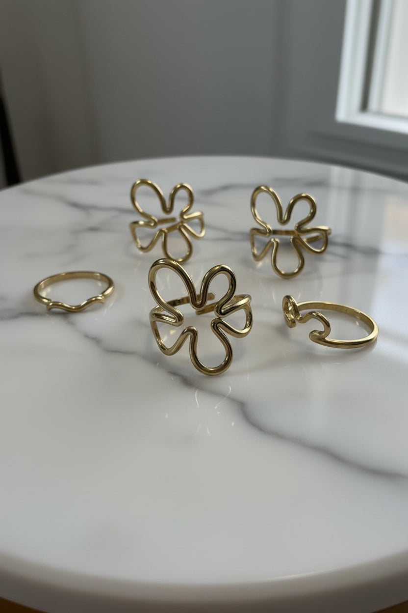 "Prachtige overzichtsfoto van de Glow Mystic in Style RVS ringen-collectie op een luxe marmeren oppervlak. Centraal staat de goudkleurige verstelbare Abstract Flower ring, omringd door andere bijpassende gouden ringen."