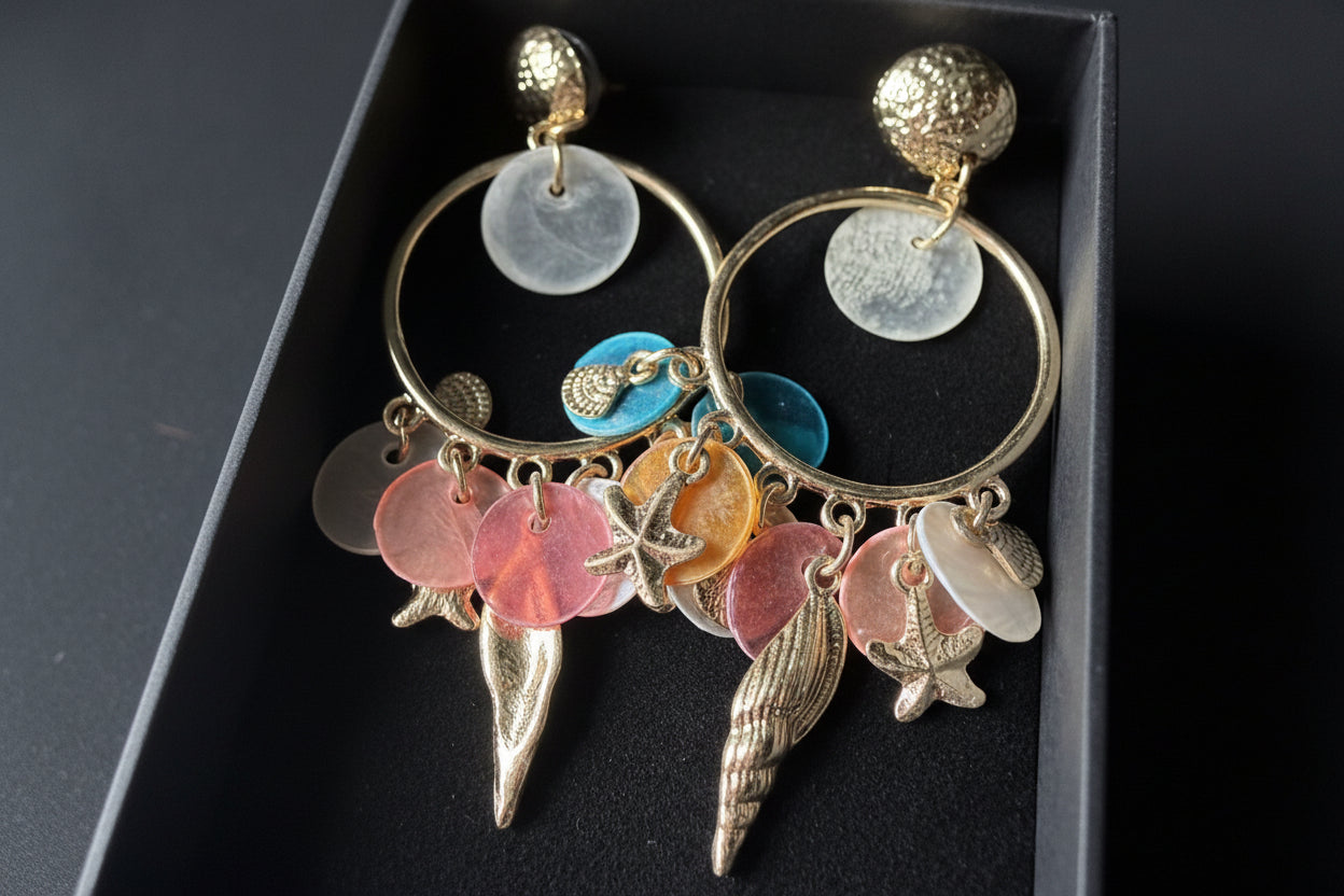 De complete Glow Mystic statement oorbellen set, gepresenteerd in een luxe zwarte velvet geschenkdoos met studio-belichting. De twee goudkleurige nikkelvrije metalen oorbellen liggen symmetrisch, waardoor alle kleurrijke charms en de gedetailleerde starfish- en conch-shellbedels goed zichtbaar zijn.