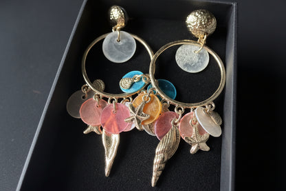 De complete Glow Mystic statement oorbellen set, gepresenteerd in een luxe zwarte velvet geschenkdoos met studio-belichting. De twee goudkleurige nikkelvrije metalen oorbellen liggen symmetrisch, waardoor alle kleurrijke charms en de gedetailleerde starfish- en conch-shellbedels goed zichtbaar zijn.