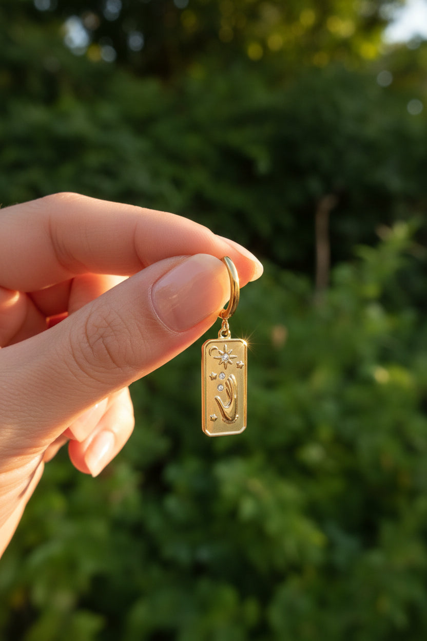Een close-up sfeerfoto van een goudkleurige RVS mystic hand oorbel, vastgehouden in een hand tegen een achtergrond van zonnige groene bladeren.