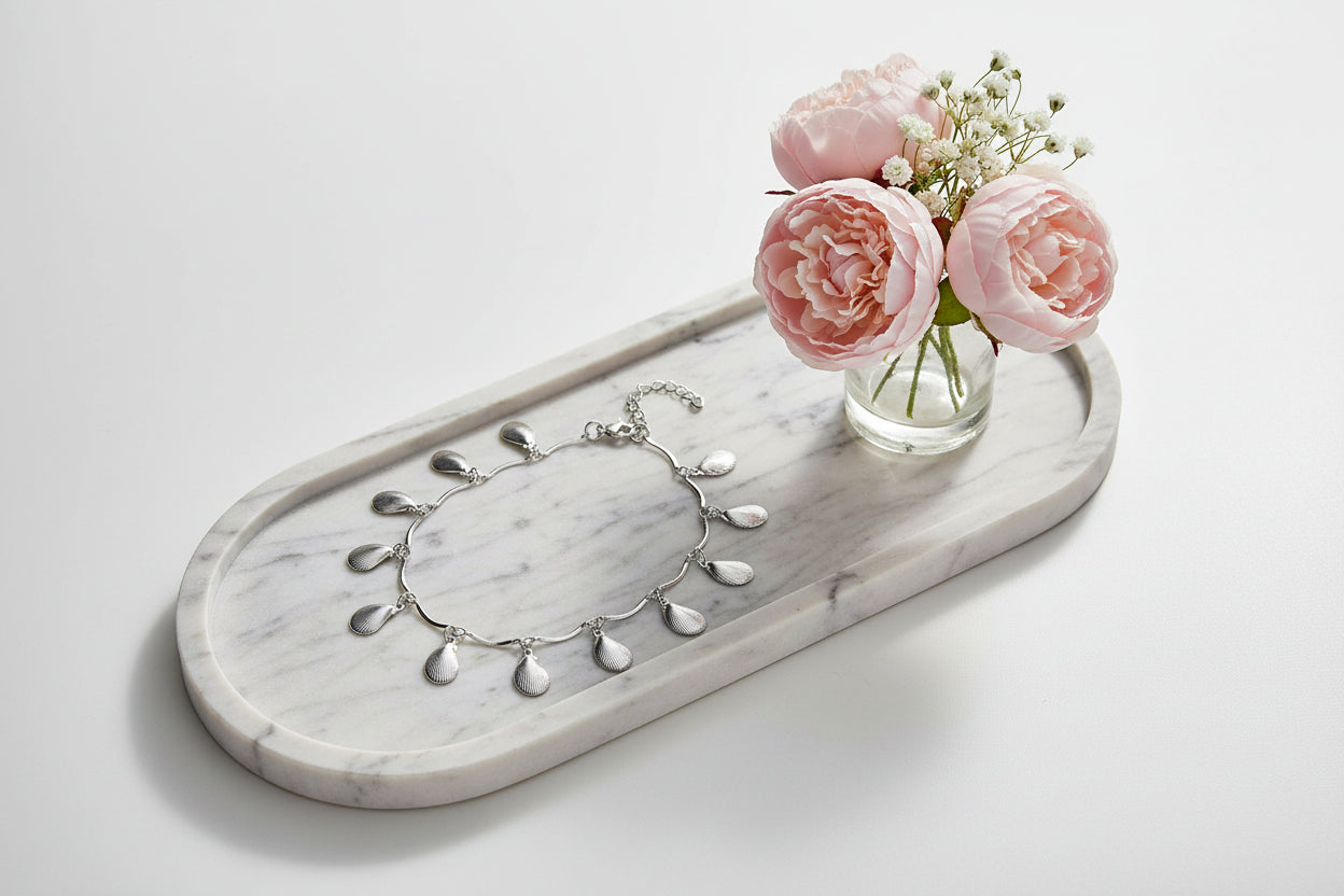 Zilverkleurig enkelbandje met hangende druppel bedels, elegant gepresenteerd op een marmeren dienblad naast een vaasje met roze bloemen