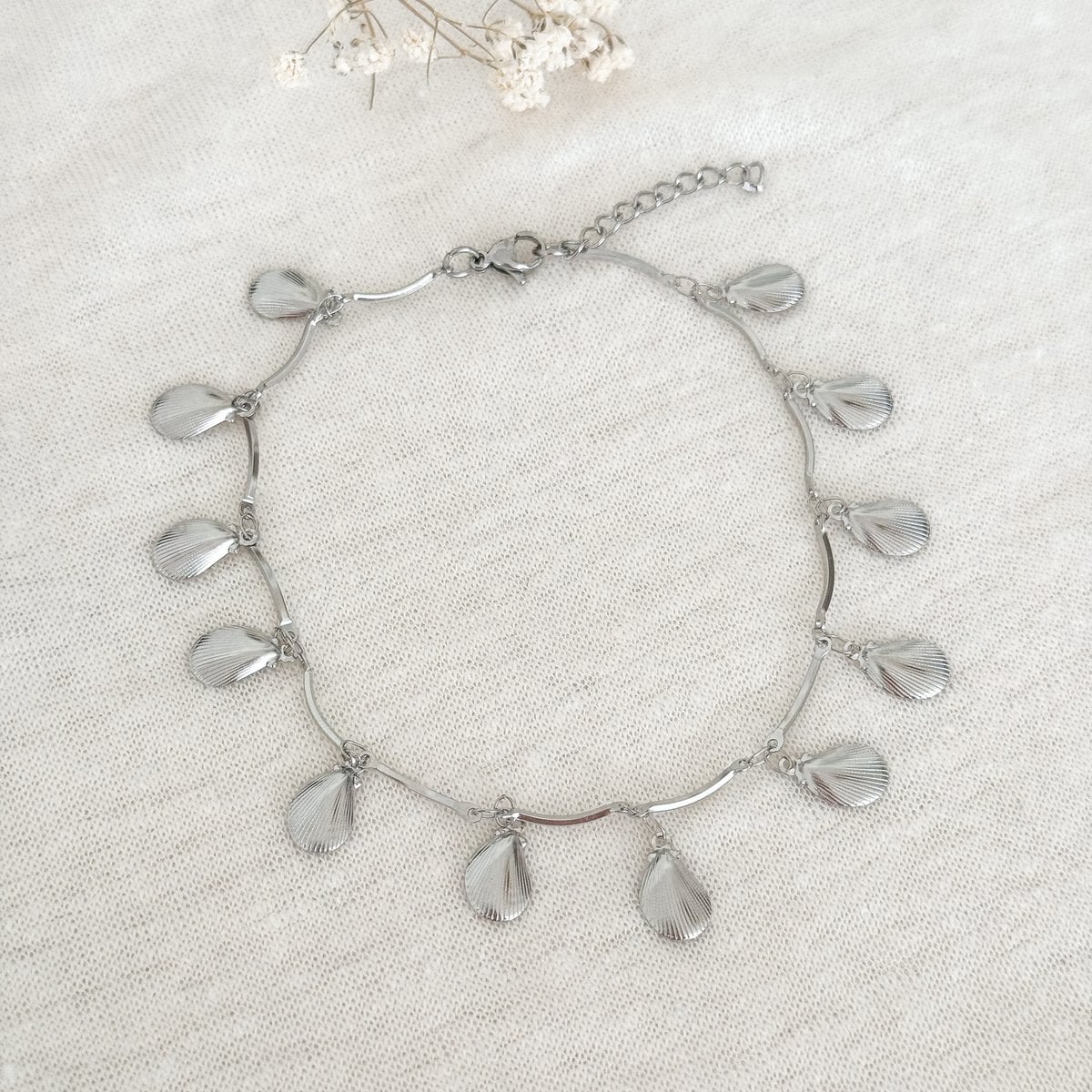 Zilverkleurige schelp enkelbandje met speelse hangertjes op een beige stoffen achtergrond met een witte droogbloem.