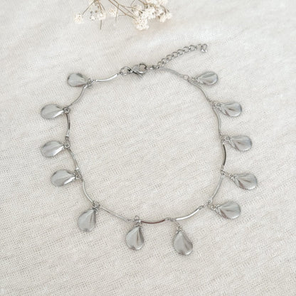 Zilverkleurige schelp enkelbandje met speelse hangertjes op een beige stoffen achtergrond met een witte droogbloem.