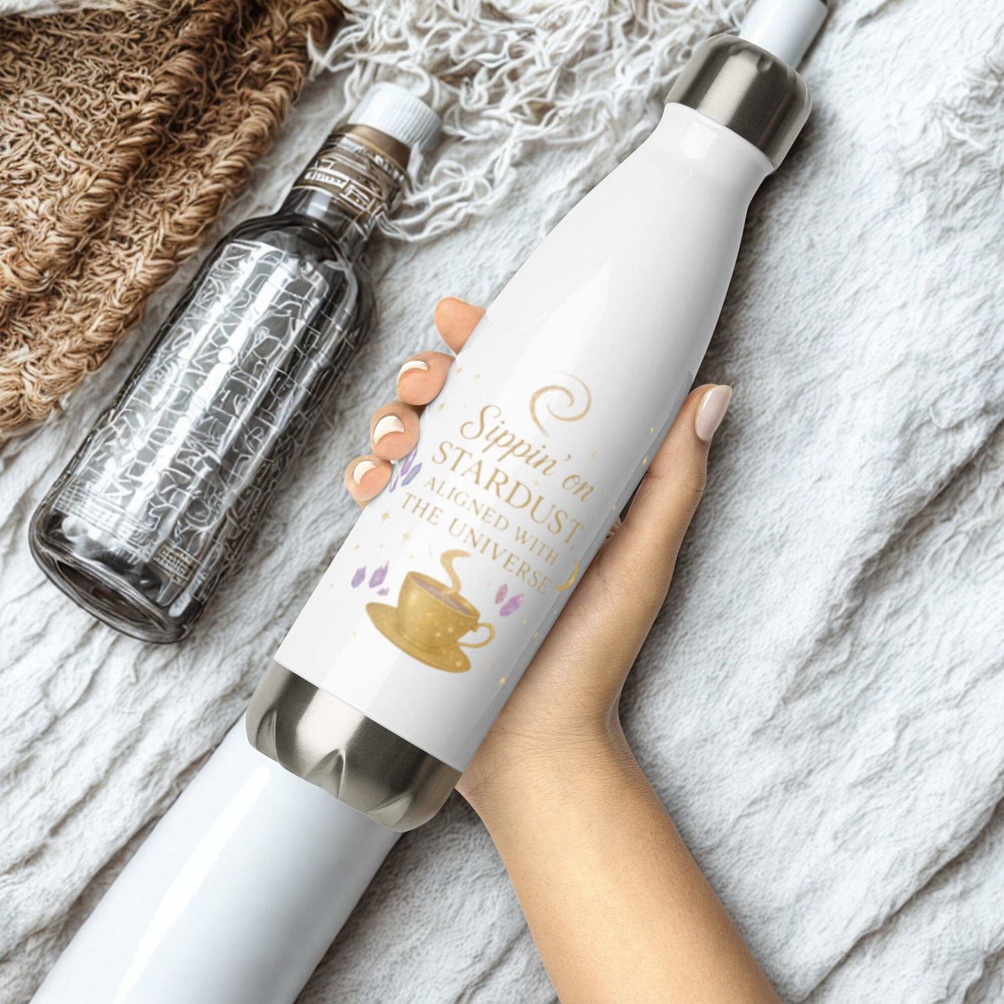 White Sippin on Stardust Water Bottle with gold text and coffee cup design held in hand on a soft textured fabric background next to a clear water bottle.