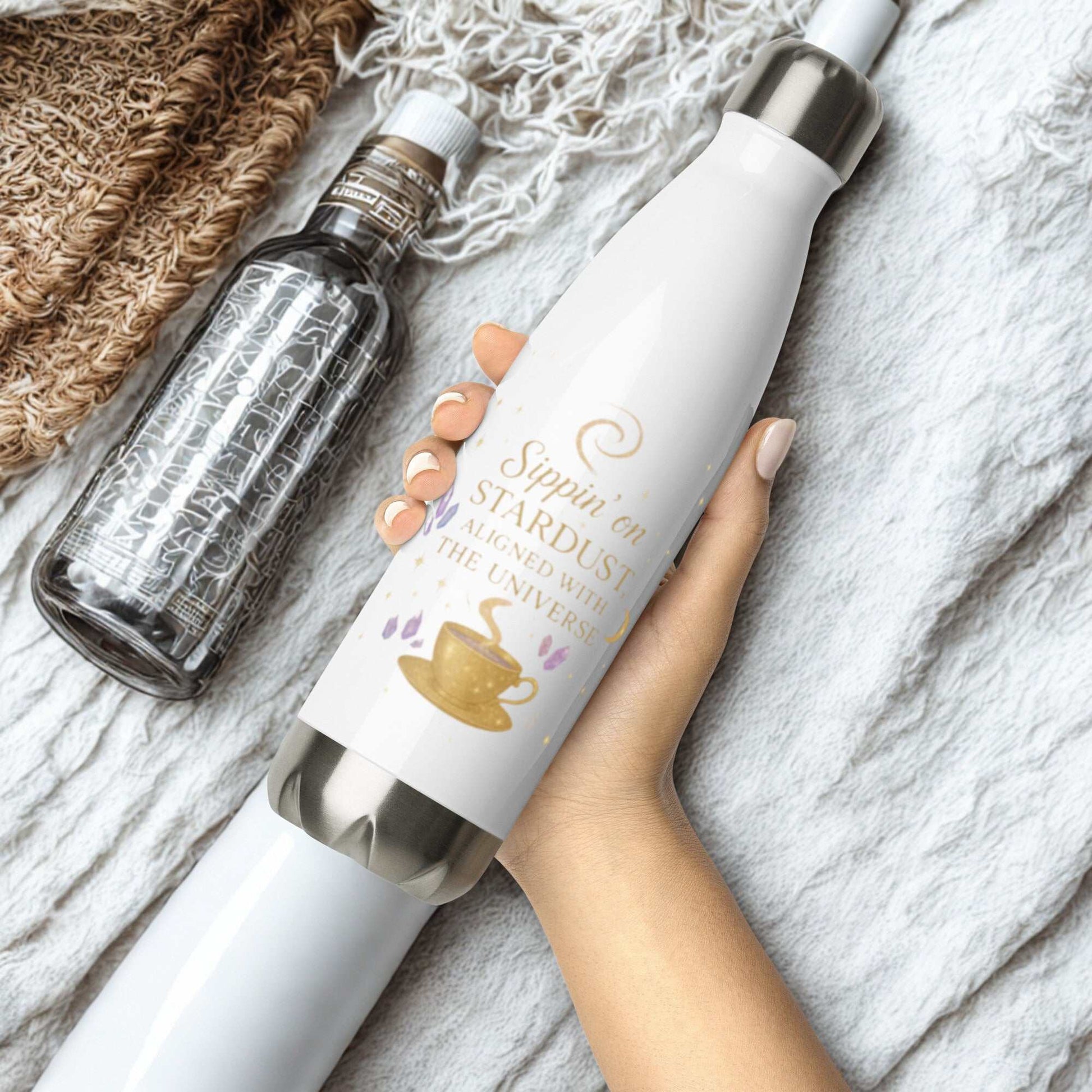 White Sippin on Stardust Water Bottle with gold text and coffee cup design held in hand on a soft textured fabric background next to a clear water bottle.