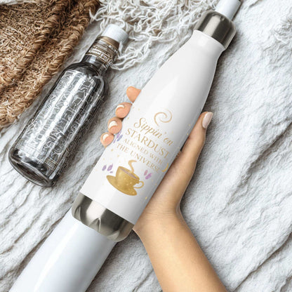 White Sippin on Stardust Water Bottle with gold text and coffee cup design held in hand on a soft textured fabric background next to a clear water bottle.